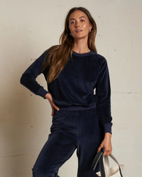 A woman in the perfectwhitetee Stella Velour Shrunken Raglan SS in Navy stands by a light wall, holding a beige and black tote bag; her relaxed look radiates casual sophistication.