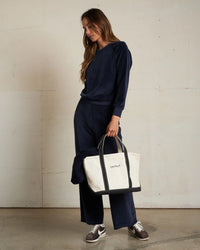 A woman wears the perfectwhitetee Stella Velour Shrunken Raglan SS in Navy with wide-leg pants indoors, carries a large white and black "perfect" tote, completes the look with brown sneakers, and lets her long hair loose.