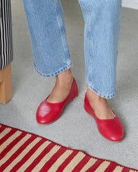 A person stands on a gray floor in beek.. Swiftlet in Lipstick blue jeans and red ballet flats with memory foam, next to a red and beige striped rug and a black and white striped chair.