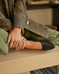 A person wearing a houndstooth coat, green pants, and beek.. Swoop in Black pointed ballet flats with memory foam sits on a wooden surface, resting their hands on their ankle and displaying rings.