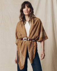A woman with wavy brown hair wears The Big Bandana in Marigold by the Great as a shawl over a white top, belted at the waist with a silver buckle and paired with dark jeans, standing against a beige background.