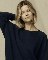 A woman with shoulder-length blonde hair wears the Great's College Sweatshirt in True Navy, standing against a neutral background with a calm, relaxed expression and one hand raised behind her head.