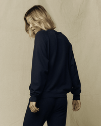 A person with shoulder-length blonde hair wears the Great’s The College Sweatshirt in True Navy, paired with relaxed-fit pants, standing against a plain beige background, turned slightly away and glancing down over their shoulder.