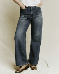 A person wearing the Great’s Dock Jean in Marina Wash, styled with a white top and brown shoes, stands against a neutral background with hands in pockets, channeling effortless 70s denim style.