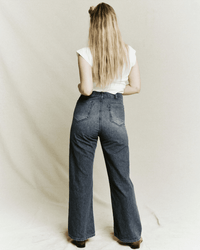 A person with long blonde hair, wearing the Great's white short-sleeve top and The Dock Jean in Marina Wash, stands facing away from the camera against a plain light backdrop.