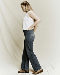 A woman with long blonde hair stands in profile against a neutral background, wearing the Dock Jean in Marina Wash by the Great, paired with a sleeveless white top and brown shoes, one hand tucked in her back pocket.