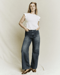 A woman stands against a plain background, wearing The Dock Jean in Marina Wash by the Great, styled with a white sleeveless top and brown shoes. She has one hand in her pocket and looks confidently at the camera.