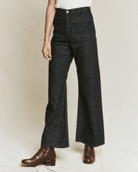 A person wearing high-waisted The Dock Jean in Rinse Wash by the Great, a white top, and brown ankle boots stands against a plain light background, with only their lower body visible.