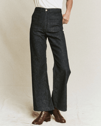 A person wears the Dock Jean in Rinse Wash by the Great—a high-waisted, wide-leg sailor jean with front patch pockets—paired with a white tucked-in shirt and brown ankle boots, standing against a plain light background.