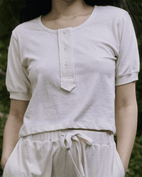A person wearing The Scoop Henley in Natural by the Great, a cream-colored 100% cotton short-sleeve buttoned shirt, paired with matching drawstring pants, stands outdoors amid greenery.