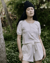 A woman wearing a black beanie and the Great's The Scoop Henley in Natural with matching shorts stands outdoors in a lush, green, wooded area.