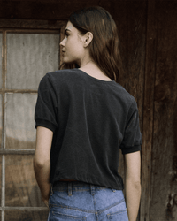 A woman with long brown hair stands outside, facing away, wearing The Scoop Henley in Washed Black by the Great with blue jeans, in front of a rustic wooden wall and window.