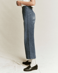 A person stands sideways in the Great’s Seafair Jean in Marina Wash, styled with a white top, black loafers, and white socks with black stripes against a plain, neutral background.