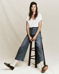 A woman with long brown hair sits on a black stool against a plain background, wearing a white tee and the Great’s Seafair Jean in Marina Wash—an ultra high-rise, wide leg style—paired with white socks with black stripes and black loafers.