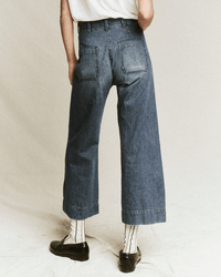 A person wearing the Great’s Seafair Jean in Marina Wash, featuring a wide-leg fit and visible back pockets, paired with a white T-shirt, striped socks, and black loafers; standing on a plain light background facing away from the camera.