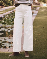 Someone wearing the Seafair Jean in True White by the Great, brown shoes, and a striped top stands on a grassy path next to a pond with lily pads in a garden setting.