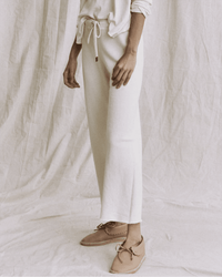 A person stands against a cream-colored fabric backdrop, wearing the Great’s Wide Leg Cropped Sweatpant in Washed White with a matching long-sleeve top and light brown moccasin-style shoes.