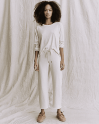 A person with curly hair stands against a light fabric backdrop, wearing a long-sleeve cream top, light brown shoes, and the Great's Wide Leg Cropped Sweatpant in Washed White.