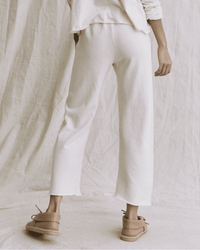 A person stands with their back to the camera, wearing the Great’s Wide Leg Cropped Sweatpant in Washed White with a matching long-sleeve top and tan shoes, set against a backdrop of soft, light-colored fabric.