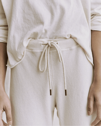 A person wears the Great's Wide Leg Cropped Sweatpant in Washed White with a loose, off-white long-sleeve shirt. Their hands relax by their sides, and the focus is on the midsection, highlighting the casual outfit.