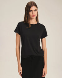 A woman with long brown hair wears the Tia Short Sleeve Crew Neck Top in Black by Velvet by Graham & Spencer, paired with a matching black skirt, standing against a neutral, light-colored background.
