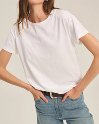 Wearing the Velvet by Graham & Spencer Tia Short Sleeve Crew Neck Top in white, styled with light blue jeans and a brown belt, this person stands with hands in pockets against a neutral background. Their head is partially out of frame.