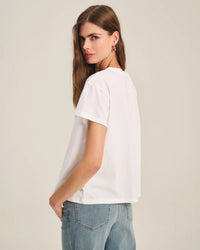 A woman with long brown hair, wearing the Velvet by Graham & Spencer Tia Short Sleeve Crew Neck Top in White and light blue jeans, stands turned slightly away from the camera, looking over her shoulder against a plain background.