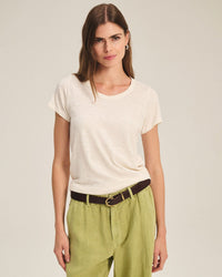 A woman with long brown hair wears a Velvet by Graham & Spencer Tilly Short Sleeve Crew Neck Top in Coconut, tucked into light green pants with a dark brown belt, standing against a plain light background.