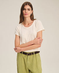 A woman with long brown hair wears the Tilly Short Sleeve Crew Neck Top in Coconut by Velvet by Graham & Spencer, paired with light green pants and a black belt, standing against a plain light background with her arms crossed.
