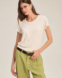 A woman with long brown hair stands against a plain background, wearing the Tilly Short Sleeve Crew Neck Top in Coconut by Velvet by Graham & Spencer, tucked into light green pants with a dark brown belt. She looks slightly to the side.