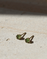 August Birthstone Studs in 14K Gold Fill by Token Jewelry—hypoallergenic gold stud earrings with round green gemstones, shown on a textured light surface in bright sunlight with shadows above.