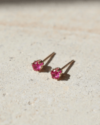 The July Birthstone Studs in 14K Gold Fill by Token Jewelry feature small round bright pink gemstones set in 14k gold fill, displayed on a light textured surface under sunlight.