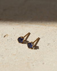 The September Birthstone Studs in 14K Gold Fill by Token Jewelry feature deep blue gemstones in handmade gold settings, perfect as birthstone studs, shown on a textured beige surface in natural sunlight.