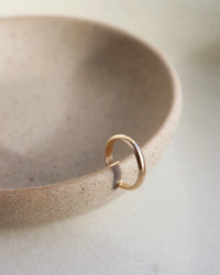 The Simple Ear Cuff 14K Gold Fill by Token Jewelry sits atop a round, beige ceramic bowl with a smooth, matte finish, its subtle elegance accentuated by soft, natural light on a pale surface.