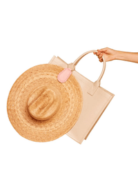 A hand holds a beige tote with a pink TopTote magnetic hat clip, The Fray in Pink, securing a wide-brimmed straw hat to the handle against a white background.