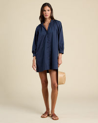 A woman stands against a plain background wearing the Angeline Dress in Navy by Trovata Birds of Paradis, a loose-fitting, long-sleeve shirtdress with a mandarin collar, crafted from 100% cotton. She pairs it with brown sandals and holds a straw hat.