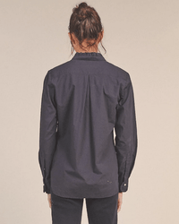 A woman with brown hair in a messy bun is seen from behind, wearing a long-sleeved Breezy Blouse in Black by Trovata Birds of Paradis and dark pants, standing against a plain light background.