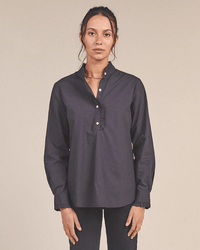 A woman with dark hair tied back stands against a plain background, wearing the Trovata Birds of Paradis Breezy Blouse in Black—a long-sleeve, 100% cotton blouse with ruffled collar and cuffs—paired with dark pants as she looks at the camera.