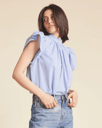 A woman with shoulder-length dark hair wears the Trovata Birds of Paradis Carla Highneck Shirt in Blue/White Stripe, featuring a ruffle collar, and high-waisted jeans. She faces sideways, looking over her shoulder with a neutral expression.