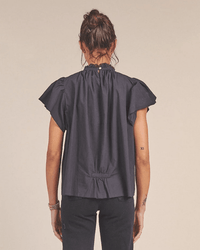 A woman with her hair in a bun stands with her back to the camera, wearing the Trovata Birds of Paradis Carla Highneck Shirt in Classic Black and black pants. A small tattoo is visible on her left arm.