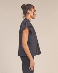 A woman with dark hair in a bun stands in profile against a plain background, wearing the Trovata Birds of Paradis Carla Highneck Shirt in Classic Black and loose black pants.