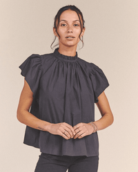 A woman faces the camera wearing the Carla Highneck Shirt in Classic Black by Trovata Birds of Paradis. The short-sleeved blouse features a high ruffle collar. Her hair is tied back with loose strands, and she holds her hands together in front.