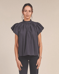 A woman faces forward, wearing the Carla Highneck Shirt in Classic Black by Trovata Birds of Paradis. The shirt features a ruffle collar and short ruffled sleeves, paired with dark pants. Her hair is tied back as she poses against a plain background.
