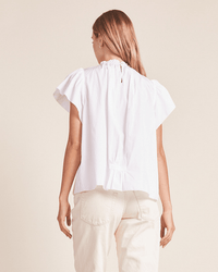 A woman with long brown hair stands facing away, wearing the Trovata Birds of Paradis Carla Highneck Shirt in White with ruffle collar and light-colored pants against a neutral background.