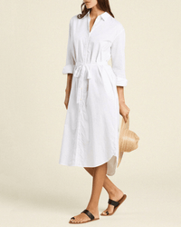 A woman wears the Catalina Shirtdress in White by Trovata Birds of Paradis, a long-sleeve, midi cotton twill dress with waist tie, paired with black sandals and holding a straw hat. The background is plain and neutral.