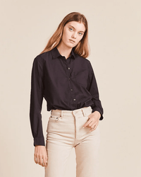 A woman with long, light brown hair wears the Trovata Birds of Paradis Grace Classic Shirt in Black, tucked into high-waisted, light beige pants, standing against a plain beige background.