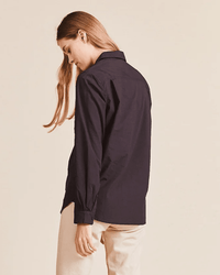 A person with long, light brown hair stands sideways and looks down, wearing a black Trovata Birds of Paradis Grace Classic Shirt and light beige pants against a plain, light background.