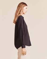A woman with long, light brown hair stands in profile against a beige background, wearing Trovata Birds of Paradis' Grace Classic Shirt in Black—crafted from 100% cotton—and light-colored pants.