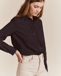 A woman with long brown hair wears the Trovata Birds of Paradis Grace Classic Shirt in Black, made from 100% cotton, tucked into high-waisted light jeans. She looks downward with one hand in her pocket against a neutral background.