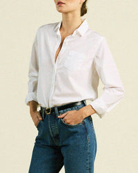 A person in a Trovata Birds of Paradis Grace Classic Shirt in White, blue jeans, and a black belt stands with hands in pockets against a plain light background. The person's head is partially out of frame.